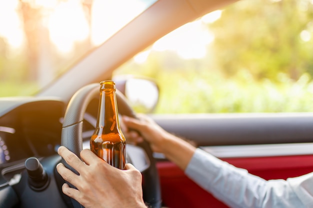 Drunk asian young man drives a car with a bottle of beer with sunset background