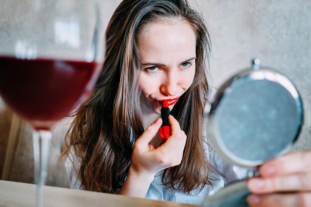 Фото Пьяная девушка с бокалом вина красит губы красной помадой.