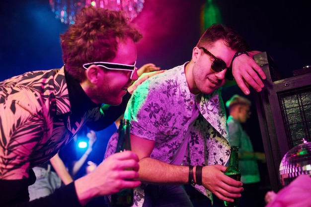 Drunk guy leaning in fence in front of young people that having fun in night club with colorful laser lights