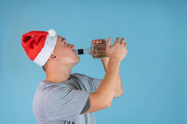 A drunk man in a red Santa hat drinks strong alcohol from the throat of a bottle standing on a blue background, copyspace