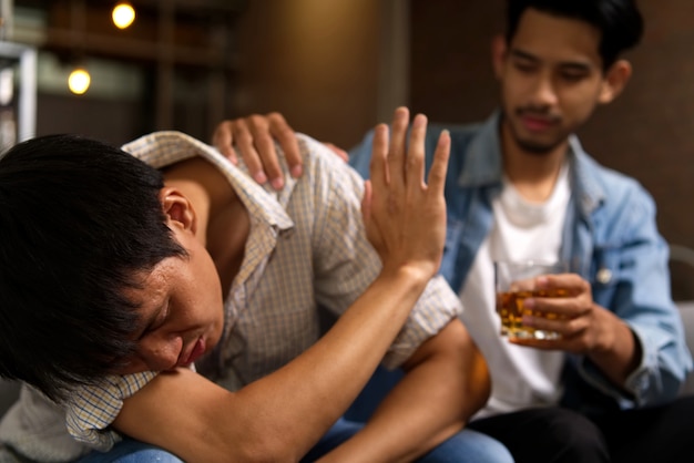 Drunk man sitting on sofa refusing whiskey from his friend by raising his hand to stop.