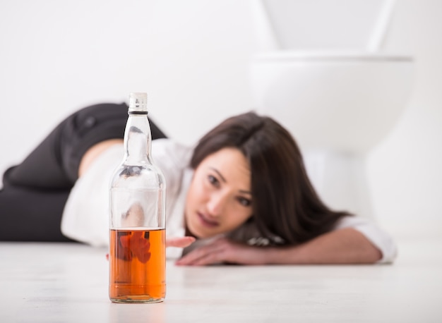 Drunk woman is lying on toilet floor with bottle of alcohol.