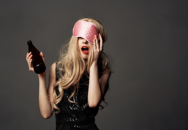 Drunk woman with a bottle of alcohol in her hand on a gray background and a mask for sleeping High quality photo