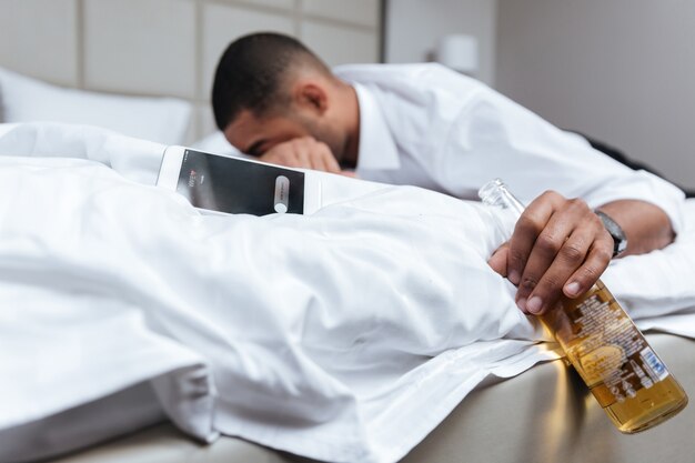 Giovane africano ubriaco in camicia che dorme sul letto e che tiene in mano la bottiglia di birra. concentrarsi sulla bottiglia. vista laterale