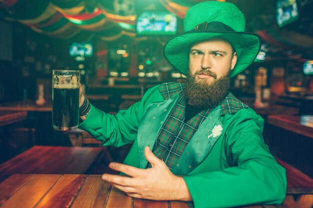 Drunk young man in green St. Patrick's suit sit at table in pub. He hold mug of dark beer. Guy chilling.