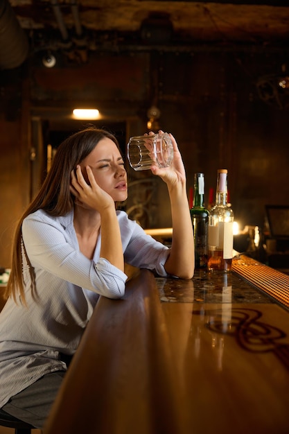 술에 취한 젊은 여성이 술집 카운터에 앉아 빈 유리를 쳐다보고 있습니다. 알코올 중독으로 고통받는 걱정스러운 사려 깊은 여성은 우울증과 번아웃을 느 ⁇ 니다.