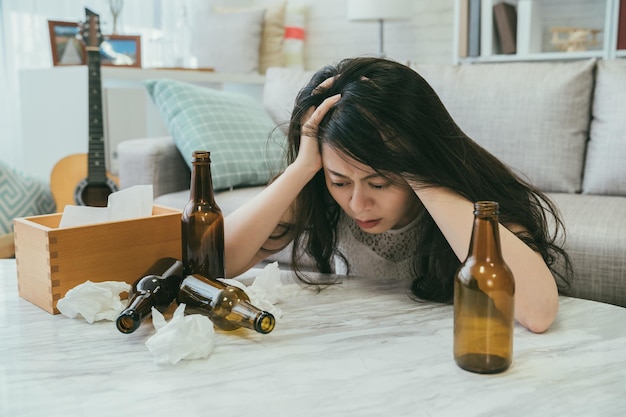 うつ病に苦しんで酔っ払ったアジアの女性は、自宅の居間で飲んだ後、崩れたティッシュとビール瓶でテーブルに頭を抱えて支えています