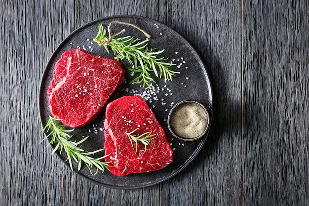 Dry-aged beef steaks with rosemary and finely ground black peppercorn on a black stone plate, horizontal view from above, flat lay, free space