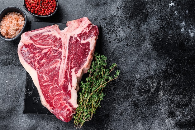 Dry-aged Raw T-bone or porterhouse beef meat Steak with herbs and salt. Black background. Top view. Copy space.