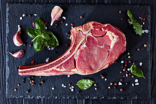 Tomahawk crudo invecchiato a secco o bistecca di manzo cowboy su una tavola di pietra con spezie, pepe in grani, aglio e basilico, close-up, piatto laico, vista da sopra