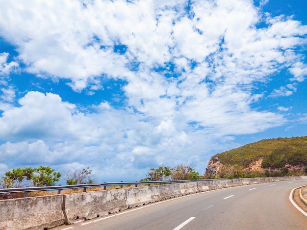 Сухая асфальтовая пустая автомобильная изогнутая дорога с бетонным барьером и линиями разметки в горах в солнечный летний день с голубым облачным небом Концепция путешествия на автомобиле