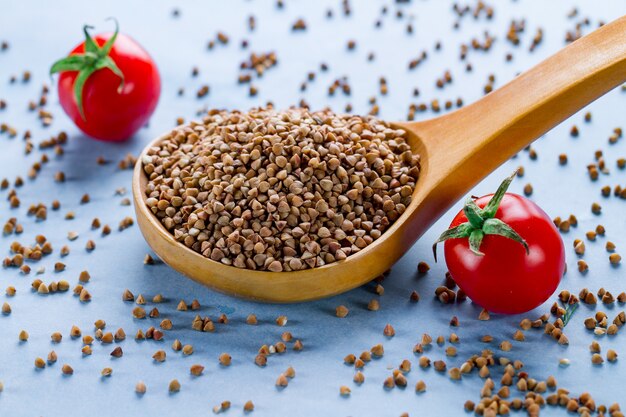 Dry buckwheat in wooden spoon and fresh ripe tomatoes cherry on blue background