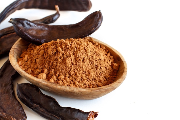 Dry carob flour and pods in a wooden bowl close up isolated on white