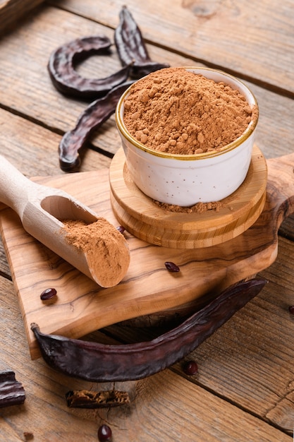 Dry carob pods and carob powder in a bowl over wooden background