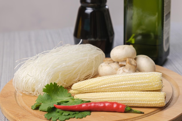 Dry funchose and ingredients for preparing an Asian dish. Glass noodles, mushrooms, mini corn, hot red pepper pod and cilantro on a wooden board. Preparing to cook Chinese noodles. Vegetarian meals.