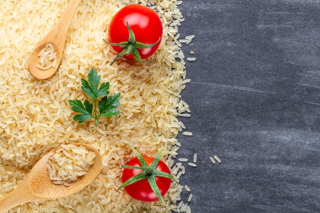 Dry golden yellow long rice with wooden spoons, ripe tomatoes cherry and fresh green parsley on dark surfce. Copy space