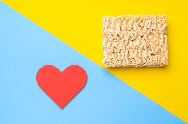 Dry instant noodles and red heart on yellow and blue background