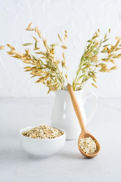 Dry oatmeal in a bowl and in a wooden spoon and a jug with ears on a light table Homemade healthy diet Vertical view