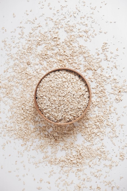 Dry oatmeal in a wooden bowl and is randomly scattered on a white background