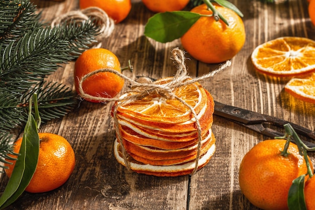 Dry oranges slices snack. Dried citrus fruits for Christmas decorations. Tangerines with leaves, fir tree branches, scissors, and thread. Wooden background, rustic concept, close up