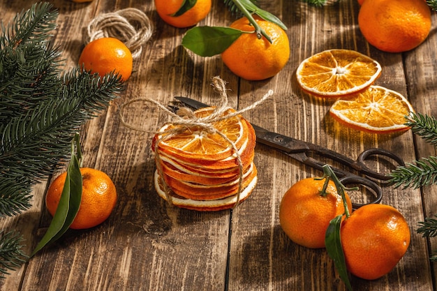 Dry oranges slices snack. Dried citrus fruits for Christmas decorations. Tangerines with leaves, fir tree branches, scissors, and thread. Wooden background, rustic concept, copy space