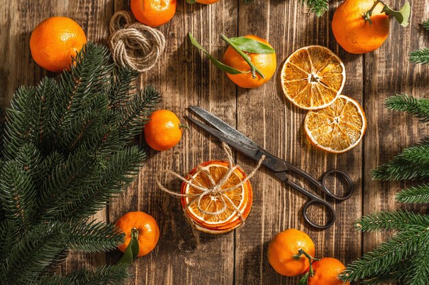 Dry oranges slices snack. Dried citrus fruits for Christmas decorations. Tangerines with leaves, fir tree branches, scissors, and thread. Wooden background, rustic concept, top view