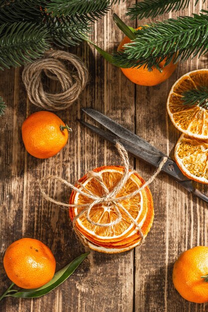 Dry oranges slices snack. Dried citrus fruits for Christmas decorations. Tangerines with leaves, fir tree branches, scissors, and thread. Wooden background, rustic concept, top view