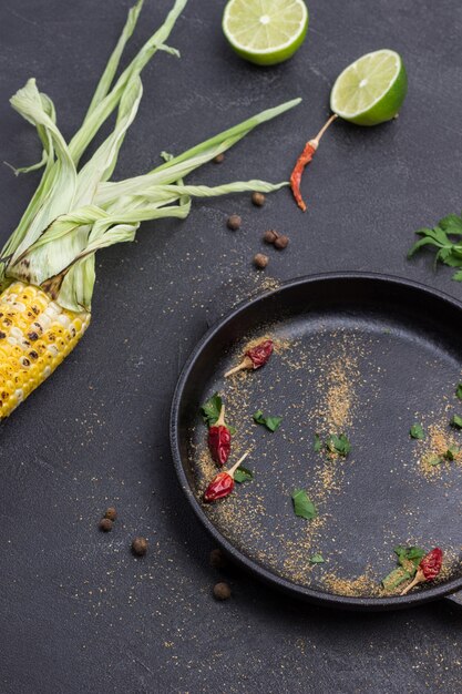 Dry red pods, ground coriander and parsley leaves in frying pan. Grilled corn and two lime halves on table. Black background. Top view