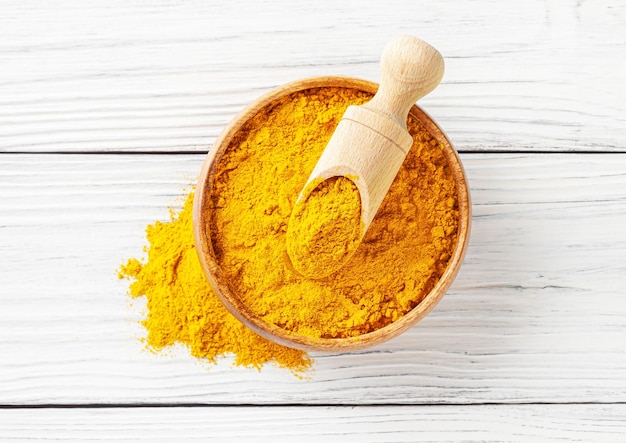 Dry turmeric curcuma powder in wooden bowl with scoop on white wooden background