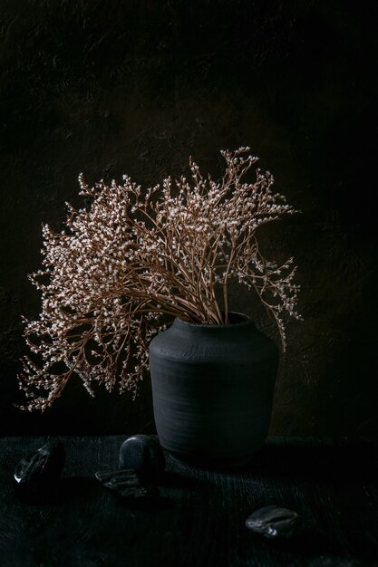 Dry white flowers branch in black ceramic vase on black wooden table with decorative stones. Dark still life. Copy space.