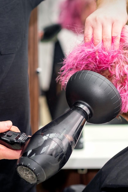 Drying short pink bob hairstyle of a young caucasian woman with\
a black hair dryer with the brush by hands of a male hairdresser in\
a hair salon close up