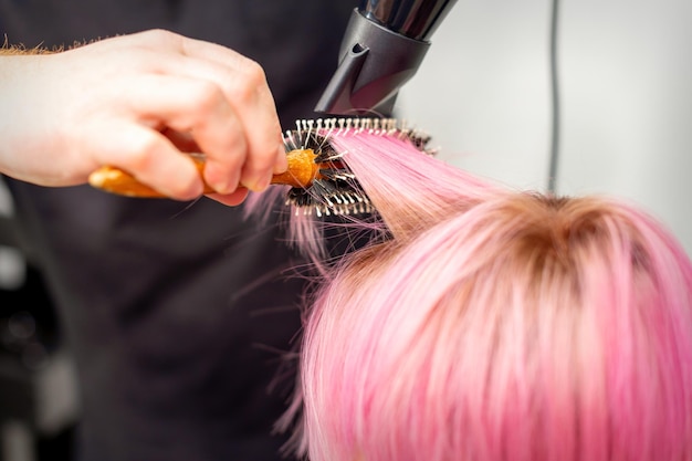 黒のヘアドライヤーと黒の丸いブラシで若い白人女性の短いピンクの髪を美容院で男性の美容師の手で乾燥させるクローズアップ