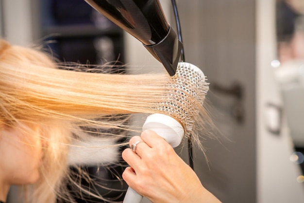 Drying straight blond hair with black hairdryer and white round brush in hairdresser salon close up