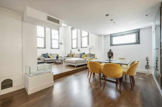 Dubbel hoog loft-appartement met vierkante eettafel met gele stoffen gestoffeerde stoelen en rondlopende fauteuils onder de ramen