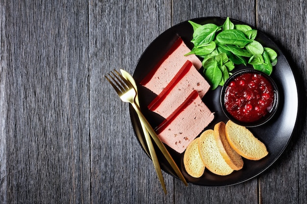 Duck liver pate with cranberry jelly on top on a black plate with cranberry sauce, spinach, and brioche croutons, with golden cutlery on a dark wooden background, close-up, top view, copy space