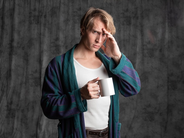 Photo dude after a party in a housecoat with a shaggy head holding a mug in his hands and suffering from a