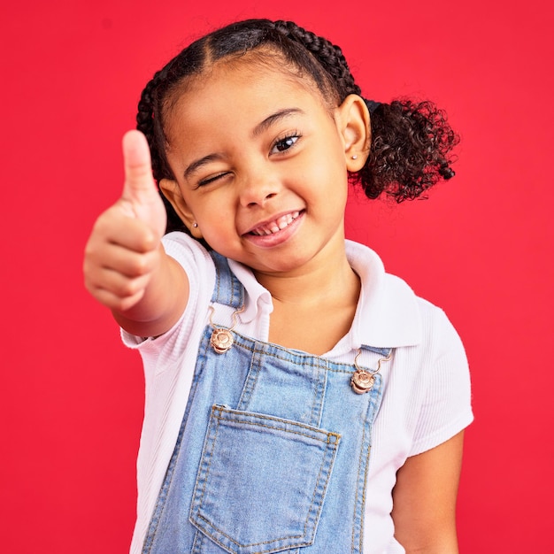 Duim omhoog portret en meisjeskind in studio rode achtergrond of geïsoleerd handteken Gelukkige kinderen duimgebaar en knipooggezicht van soortgelijke emoji-ondersteuning en blijk van vertrouwen bedankt of ja akkoord van beoordeling