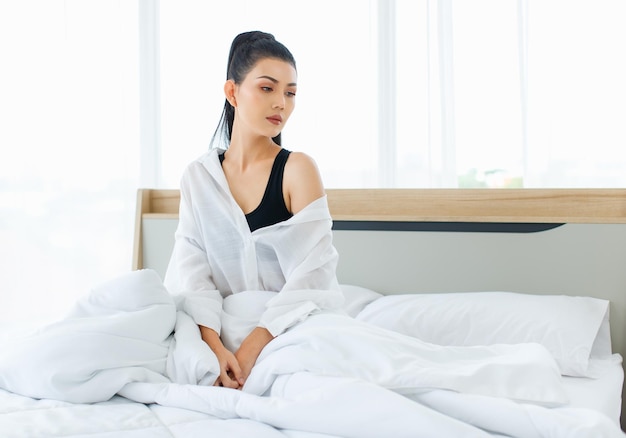 Duizendjarige Aziatische sexy doordachte vrouw in casual comfortabele pyjama zittend onder een witte dikke warme deken vanaf de dag denkend naar buiten alleen te kijken na het wakker worden in de slaapkamer in de ochtend thuis