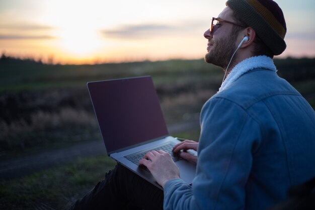 Duizendjarige hipster man aan het werk op laptop met koptelefoon in veld bij zonsondergang