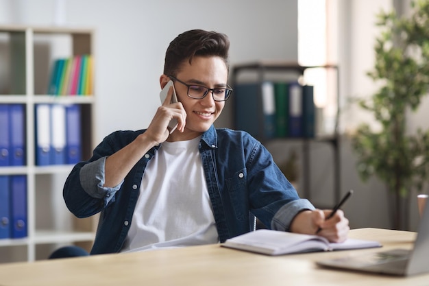 Duizendjarige man die notities schrijft tijdens het telefoneren op de werkplek op kantoor