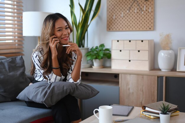 Duizendjarige vrouw freelancer zittend in de woonkamer en praten op mobiele telefoon