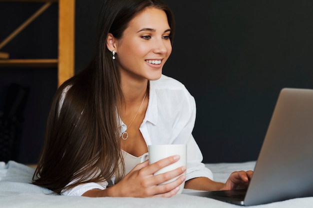 Duizendjarige vrouw zit op bed in wit overhemd, kopje koffie in de hand te houden, laptop scherm kijken. Mooie brunette vrouw gebruikt laptop om online inhoud, online training te bekijken, op afstand te werken