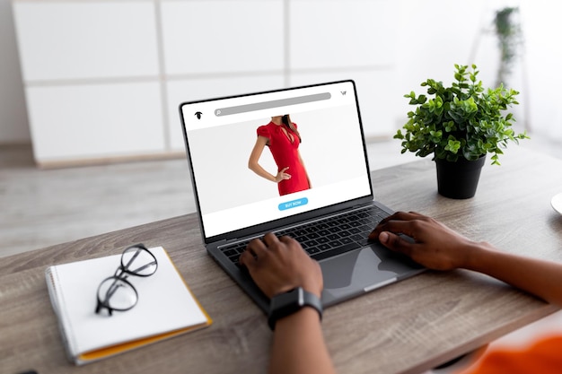 Duizendjarige zwarte vrouw met laptop die nieuwe winkelwebsite thuis of op kantoor gebruikt, kiest kleding