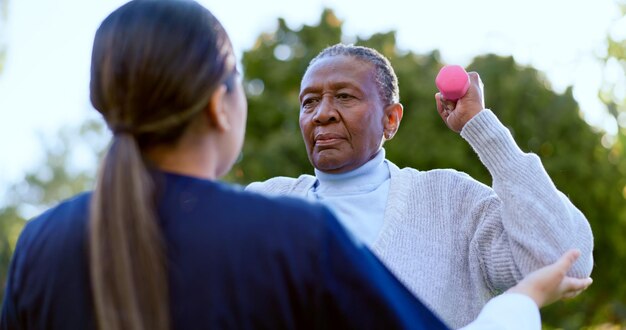 Photo dumbbell exercise and a senior black woman with a nurse outdoor in a garden together for physiotherapy fitness health or wellness with an elderly patient and medical nurse in the yard to workout