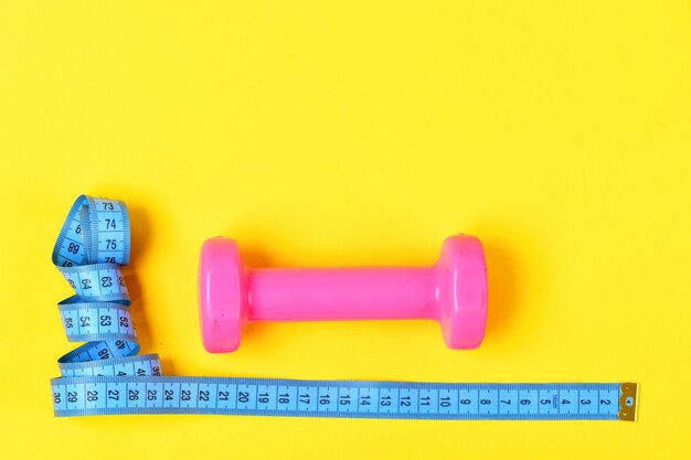 Dumbbell in pink color and measuring tape lay top on yellow background. Weight loss and sports concept. Centimeter in blue color near colorful sport equipment. Gym and healthy lifestyle tools.