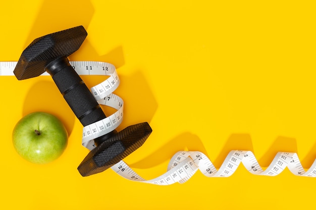 Dumbbells, apple and measure tape on yellow background. Healthy lifestyle or weight loss concept. Copy space