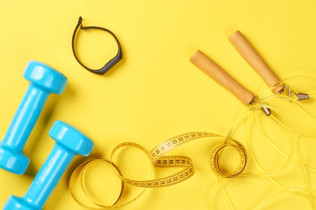 Dumbbells, fitness tracker, jump rope and measuring tape on a yellow background