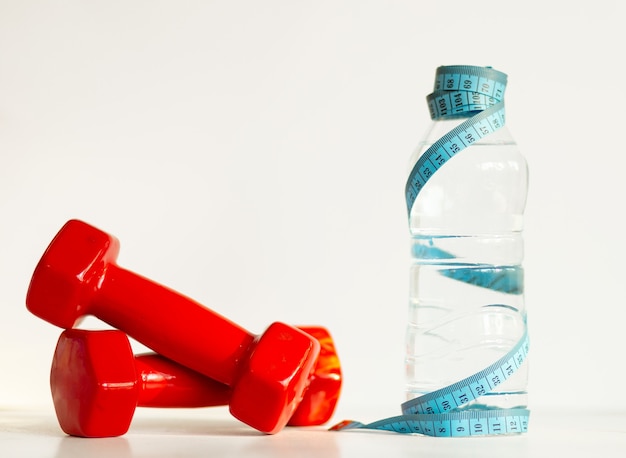 Dumbbells, glass bottle of water, fresh green apple, measuring tape on a light background. Fitness at home. Healthy lifestyle. Healthy habits. Copy space. Close up.