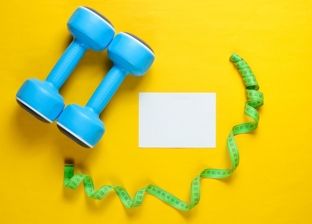 Dumbbells, measuring tape and blank card on yellow surface