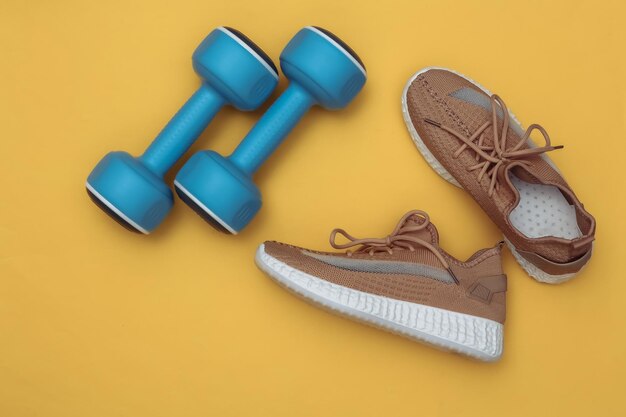 Dumbbells and textile sports shoes on yellow background. Fitness flat lay. Top view